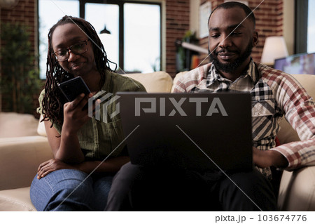African american woman browsing social media on smartphone while boyfriend is working on laptop sitting on couch. Couple spending free time together using digital devices in home living room. 103674776