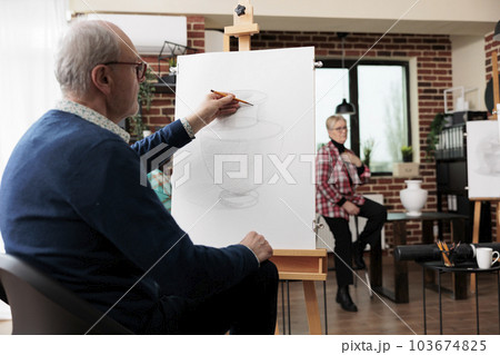 Senior man student sitting at easel using pencil drawing on canvas, active retiree learning how to draw with teacher in art group, enjoying creative activities in relaxed peaceful atmosphere 103674825