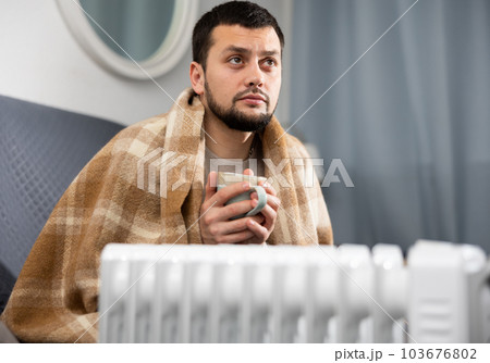 Guy wrapped in blanket sitting with cup next to heater in apartment 103676802