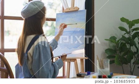 Young female painter using paintbrush to drawing and painting masterpiece on canvas in home studio 103685935