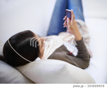 Enjoying some tunes. A young woman listening to her mp3 player while lying on the sofa. 103689959