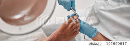 Skilled podiatrist in gloves cuts toenail of client with clipper in salon Skilled podiatrist in gloves cuts toenail of client with clipper in salon 103690617