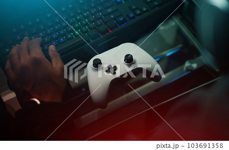Hand, video game controller and keyboard on desk, gamer setup and online streaming with competition and esports. Gaming technology, streamer person in dark room and playing tournament with joystick Hand, video game controller and keyboard on desk, gamer setup and online streaming with competition and esports. Gaming technology, streamer person in dark room and playing tournament with joystick 103691358