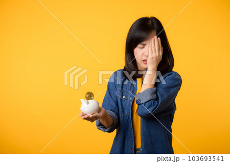 Unhappy asian young woman wearing yellow t-shirt denim shirt pulling digital coin crypto currency out of piggy bank isolated on yellow background. Payment digital money debt financial concept. Unhappy asian young woman wearing yellow t-shirt denim shirt pulling digital coin crypto currency out of piggy bank isolated on yellow background. Payment digital money debt financial concept. 103693541