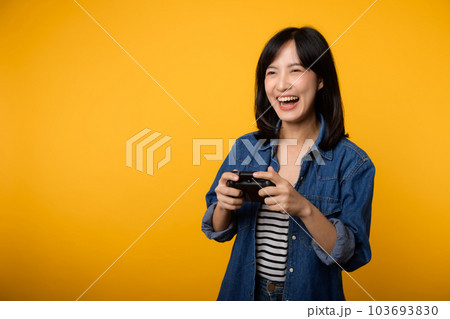 Portrait young asian woman with happy success smile wearing denim clothes holding joystick controller and playing video game. Fun and relax hobby entertainment lifestyle concept. 103693830