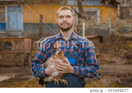 Farmer smiling European appearance male Caucasian rural portrait on countryside with a beard, shirt and overalls with a white-necked chicken in his arms outdoors Farmer smiling European appearance male Caucasian rural portrait on countryside with a beard, shirt and overalls with a white-necked chicken in his arms outdoors 103698077