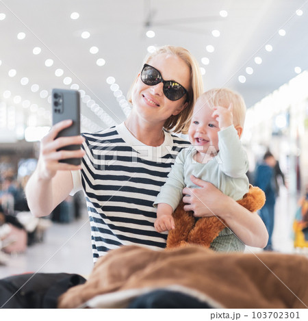 Mother taking selfie with mobile phone, while traveling with child, holding his infant baby boy at airport, waiting to board a plane. Travel with kids concept Mother taking selfie with mobile phone, while traveling with child, holding his infant baby boy at airport, waiting to board a plane. Travel with kids concept 103702301