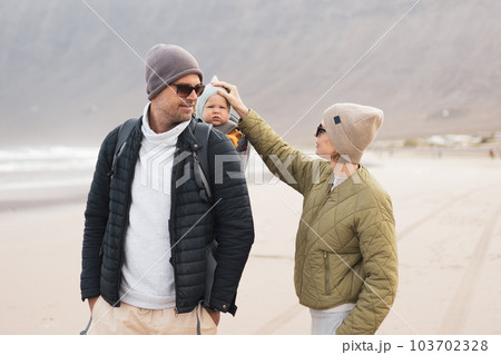 Young father carrying his infant baby boy son in backpack on windy sandy Famara beach, Lanzarote island, Spain. Family travel and winter vacation concept 103702328
