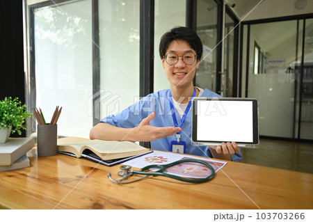 Young surgeon wearing blue scrubs holding and showing digital tablet with blank screen. Healthcare and medical concept Young surgeon wearing blue scrubs holding and showing digital tablet with blank screen. Healthcare and medical concept 103703266