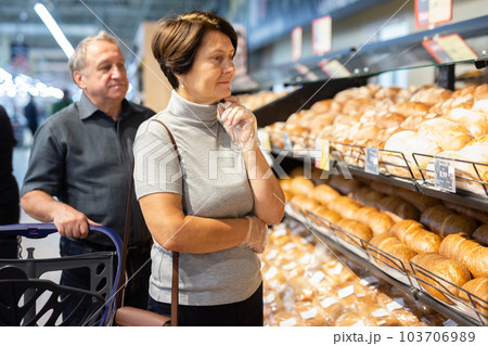 Elderly married couple chooses hot fresh buns and bread at supermarket showcase Elderly married couple chooses hot fresh buns and bread at supermarket showcase 103706989