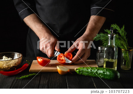 Chef hands with knife, cutting fresh red tomatoes for salad. Working environment in the restaurant kitchen. Fresh vegetables and oil on table 103707223