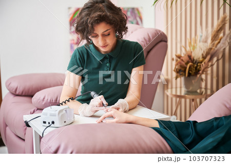 Manicurist removing cuticle on client hand with electric nail drill. Female manicure master sitting at the table while doing hardware manicure on woman hand. Manicurist removing cuticle on client hand with electric nail drill. Female manicure master sitting at the table while doing hardware manicure on woman hand. 103707323