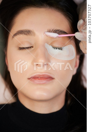 Woman with eye patch under lower eyelid keeping eyes closed while beautician applying cleansing mousse on eyebrows with lash brush. Female person having eyelash extension procedure in beauty salon. 103707327