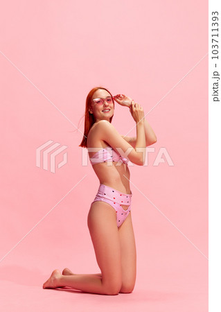 Portrait of smiling, attractive, young, redhead girl with perfect, slim body posing in swimsuit against pink studio background 103711393