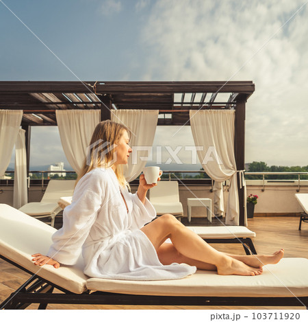 Woman in white bathrobe lying on sofa and relaxing with orange cocktail at the roof. Woman in white bathrobe lying on sofa and relaxing with orange cocktail at the roof. 103711920