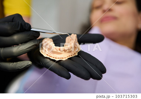 Dentist showing dental plaster mold to the camera, holding hands above a female patient lying in dentist's chair, getting treatment in dentistry clinic. Jaw model. Health, dental and oral care concept Dentist showing dental plaster mold to the camera, holding hands above a female patient lying in dentist's chair, getting treatment in dentistry clinic. Jaw model. Health, dental and oral care concept 103715303