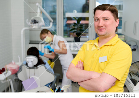 Confident portrait of a Caucasian young happy male dentist orthodontist in yellow medical uniform, standing with arms folded in the dental office in modern dentistry clinic, smiling looking at camera Confident portrait of a Caucasian young happy male dentist orthodontist in yellow medical uniform, standing with arms folded in the dental office in modern dentistry clinic, smiling looking at camera 103715313