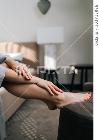 Vertical cropped shot of unrecognizable sensual naked female legs and hands, smearing cream on leg. Closeup side view of pretty young woman touching smooth skin with manicured hands sitting on bed. Vertical cropped shot of unrecognizable sensual naked female legs and hands, smearing cream on leg. Closeup side view of pretty young woman touching smooth skin with manicured hands sitting on bed. 103715926