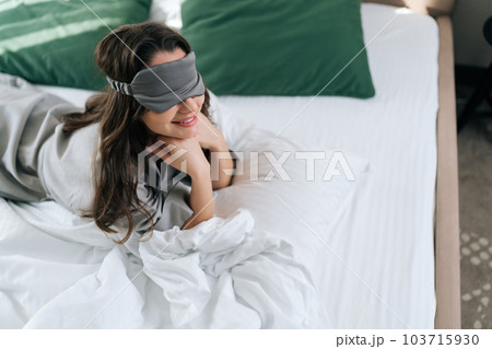 High-angle view of smiling young woman wearing sleeping mask lying in comfortable bed in luxury bedroom in hotel. Portrait of beautiful brunette female in pajama waking up enjoy good morning at home. 103715930