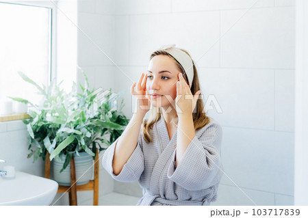 Young woman in bathrobe looking in mirror and doing manual self massage of face in the bathroom. Natural procedures for skin care. Home Beauty treatment, face gymnastics for youth. Selective focus Young woman in bathrobe looking in mirror and doing manual self massage of face in the bathroom. Natural procedures for skin care. Home Beauty treatment, face gymnastics for youth. Selective focus 103717839
