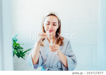 Young woman in bathrobe looking in the mirror, holding dropper with hyaluronic acid serum close to face and smiling. Skin hydrating. Cosmetic spa procedures. Beauty self-care at home. Selective focus 103717877
