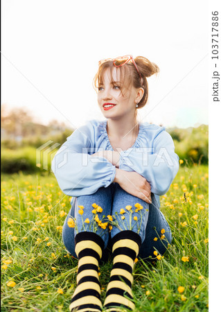 Young woman wearing blue blouse, jeans and striped black and yellow socks with flowers inside sitting on the green grass of blooming meadow. Anime style. Concept of bee protection, bloom season. Young woman wearing blue blouse, jeans and striped black and yellow socks with flowers inside sitting on the green grass of blooming meadow. Anime style. Concept of bee protection, bloom season. 103717886