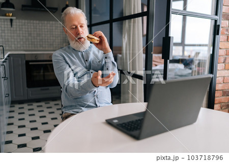 Hungry gray-haired mature elderly man eating hamburger with beef from fast food restaurant watching online cinema on laptop computer, looking to screen, sitting at table in in modern kitchen room. 103718796