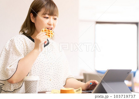 ワッフルを食べながらタブレットの画面を見る若い女性 103719737