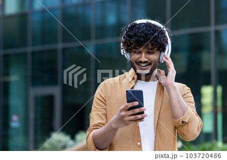 A young male Hispanic student is standing on the street near the campus wearing headphones with a phone in his hands. Listens to music, a podcast, talks on a video call. A young male Hispanic student is standing on the street near the campus wearing headphones with a phone in his hands. Listens to music, a podcast, talks on a video call. 103720486