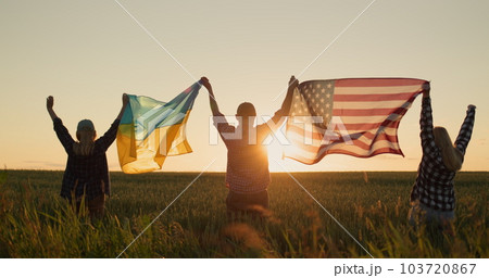 A group of friends raises the flag of the United States and the flag of Ukraine over their heads. Standing against the backdrop of a field of wheat where the sun sets 103720867