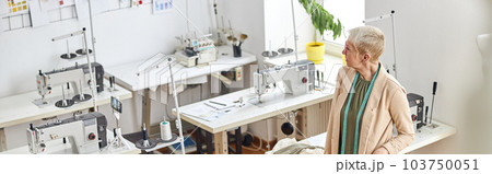 Mature dressmaker with stylish short haircut in empty sewing workshop 103750051