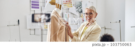 Smiling woman tailor shows new beige jacket shooting video while African-American seamstress sits at workplace Smiling woman tailor shows new beige jacket shooting video while African-American seamstress sits at workplace 103750063