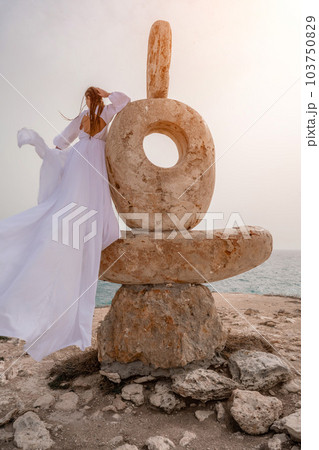 Woman sea stone. A woman stands on a stone sculpture made of large stones. She is dressed in a white long dress, against the backdrop of the sea and sky. The dress develops in the wind. 103750829