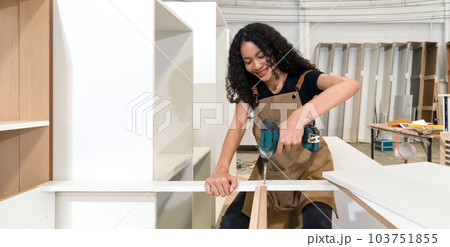 Teenager in brown apron use cordless electric drill with battery to assemble wooden furniture in the factory. Wooden cabinets of various sizes are placed in the work area. Teenager in brown apron use cordless electric drill with battery to assemble wooden furniture in the factory. Wooden cabinets of various sizes are placed in the work area. 103751855