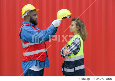 A foreman with mustache and beard helps a female apprentice wear a hardhat. Both of them wearing reflective safety vest. A large cargo container is in the background. A foreman with mustache and beard helps a female apprentice wear a hardhat. Both of them wearing reflective safety vest. A large cargo container is in the background. 103751860