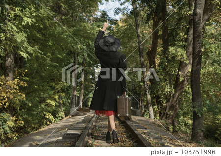 Woman in stylish coat, broad-brim hat and with vintage suitcase walks along railway tracks going through forest. Back view Woman in stylish coat, broad-brim hat and with vintage suitcase walks along railway tracks going through forest. Back view 103751996