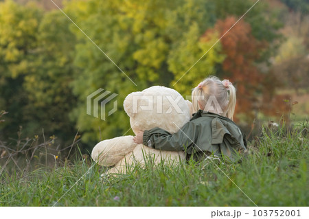 Blonde little girl and teddy bear on grass on autumn trees backgound. Child with soft toy rests in nature Blonde little girl and teddy bear on grass on autumn trees backgound. Child with soft toy rests in nature 103752001