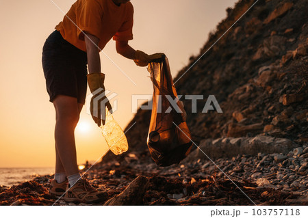 Ecology. A volunteer collecting plastic bottles on the beach in to the bag. Sunset in the background. The concept of Earth Day and environmental conservation 103757118