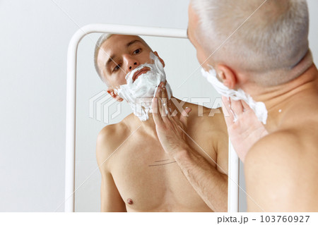 Portrait of young shirtless man shaving his beard, looking in mirror against white studio background 103760927