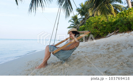 Young woman in straw hat making notes while resting in swings on tropical beach 103769418