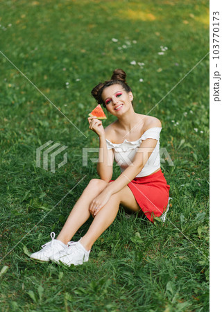 A smiling girl holds a sweet watermelon in her hands and smiles while sitting on the grass 103770173