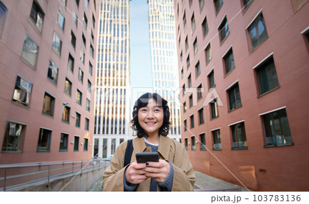 Portrait of cute girl tourist, korean woman in headphones, looks at mobile phone, uses smartphone app, map or text messages, listens music in headphones 103783136