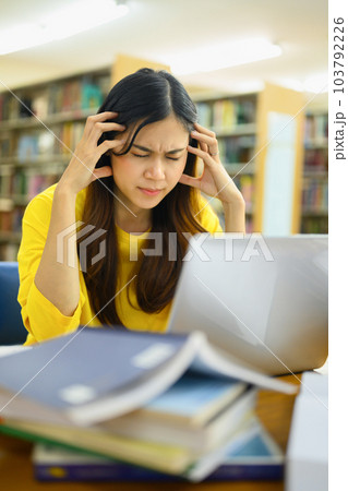 Exhausted female student sitting in library with laptop, suffering from headache or feeling tired of studying for college exam Exhausted female student sitting in library with laptop, suffering from headache or feeling tired of studying for college exam 103792226