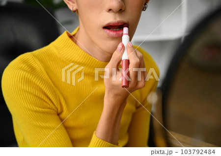 Cropped image of young gay man doing makeup and applying pink lipstick on lips in front of mirror at home Cropped image of young gay man doing makeup and applying pink lipstick on lips in front of mirror at home 103792484