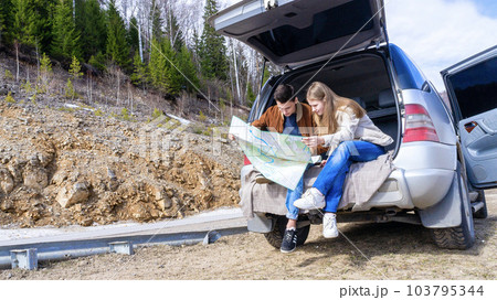 A happy young couple sits at the trunk of a car reading a road map against the backdrop of mountains. 103795344