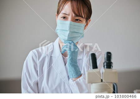 デスクで顎に手を当てて考え事をする白衣の若い女性 103800466