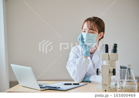 デスクで憂鬱そうな表情をする白衣の若い女性 103800469