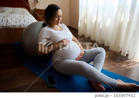 Health pregnancy, motherhood, procreation concept. Portrait of a sporty expectant loving caring mother waiting for newborn baby, holding her hands on her tummy, gently stroking it, sitting on yoga mat 103807452