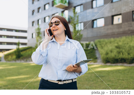 Lifestyle portrait of a stylish young businesswoman talking on cell phone, making call and working online, standing on city street. Successful business. Happy female using modern technology Lifestyle portrait of a stylish young businesswoman talking on cell phone, making call and working online, standing on city street. Successful business. Happy female using modern technology 103810572