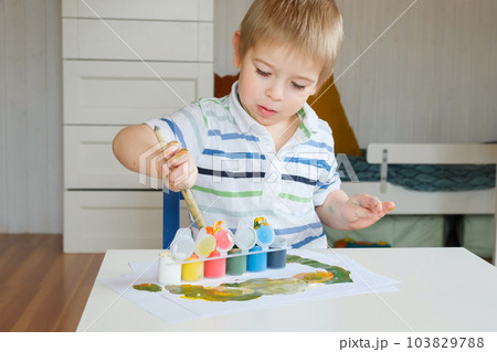 Little Boy Draws Picture On Paper. Preschool Child in striped clothes drawing with Paints at the Table at Home. Day. Real People. Creative little Kid. Little Boy Draws Picture On Paper. Preschool Child in striped clothes drawing with Paints at the Table at Home. Day. Real People. Creative little Kid. 103829788